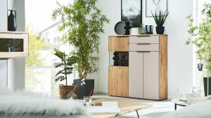 Modernes Wohnzimmer mit hellen Holzmöbeln, Zimmerpflanzen, Kunst an weißen Wänden und großen Fenstern, die natürliches Licht hereinlassen. Ein Schrank mit Dekoration und ein Tisch mit einem Getränk und einem Buch sind sichtbar. Die Atmosphäre ist hell und gemütlich.