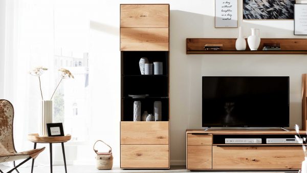 Ein modernes Wohnzimmer mit der Interliving Wohnzimmer Serie 2020 - Vitrine 151302, hellen Holzmöbeln, einem Fernseher auf einem niedrigen Schrank, Dekorationsartikeln auf hohen Regalen und Wandregalen mit Vasen und gerahmter Kunst, die durch transparente Vorhänge in das Sonnenlicht getaucht werden.