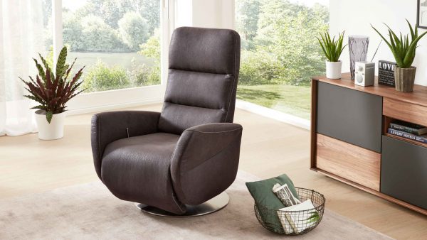 Der Interliving Sessel Serie 4530, ein moderner dunkelgrauer Drehsessel, steht auf einem hellen Teppich in einem hellen Wohnzimmer mit großen Fenstern, Grünpflanzen, einem Holzschrank und einem Drahtkorb mit einer Decke.