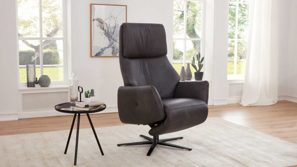 Der Interliving Sessel Serie 4520 - Sessel 77V44, ein moderner schwarzer Drehsessel, steht auf einem hellen Teppich in einem hellen Wohnzimmer mit großen Fenstern, einem Beistelltisch mit Büchern und Dekoration, einer Topfpflanze und Wandkunst.