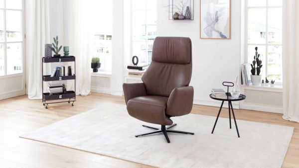In einem modernen Wohnzimmer steht der Interliving Sessel Serie 4520 - Sessel 45V44, ein brauner Lederdrehsessel auf einem hellen Teppich neben einem kleinen runden Tisch mit Büchern und einem Schlüssel. Große Fenster, Pflanzen und ein Barwagen bereichern den hellen, minimalistischen Raum.