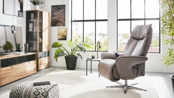 In einem modernen Wohnzimmer mit großen Fenstern stehen die Interliving Sessel Serie 4552 - Sessel 58U44 in Grau, ein Holzschrank, ein TV-Ständer, Topfpflanzen, gerahmte Wandkunst und ein gemusterter Pouf auf einem hellen Teppich. Der Raum ist von natürlichem Licht durchflutet.