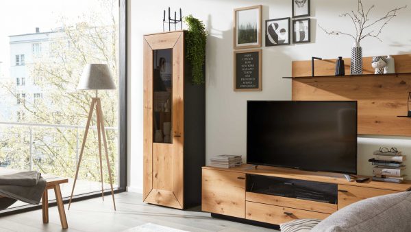 Ein modernes Wohnzimmer mit der Interliving Wohnzimmer Serie 2022 - Vitrine Z11-2L, hellen Holzmöbeln, einem Flachbildfernseher, einer Stehlampe, Wandkunst, Regalen mit Dekoration und großen Fenstern mit Blick auf Bäume.