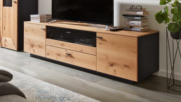 Das Interliving Wohnzimmer Serie 2022 - Lowboard LB6 ist ein moderner TV-Ständer aus Holz mit schwarzen Akzenten, geschlossenen Schränken, Schubladen und einer Glasplatte. Es bietet Platz für einen Flachbildfernseher, Bücher und Gläser auf einem grauen Holzboden neben einer grünen Pflanze.