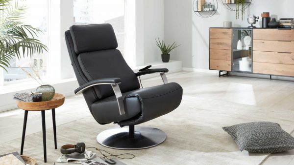 Der Interliving Sessel Serie 4541 - Relaxsessel F, ein moderner schwarzer Ruhesessel mit Metallarmlehnen, steht auf einem beigen Teppich in einem hellen Wohnzimmer mit hellem Holzboden, Topfpflanzen, einem Holzsideboard und verschiedenen Accessoires.