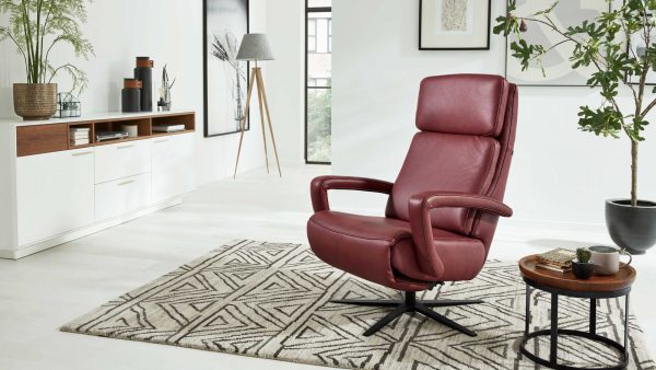 Ein modernes Wohnzimmer mit dem Interliving Sessel Serie 4541 - Relaxsessel C in burgunderrotem Leder auf einem geometrischen Teppich, gepaart mit einem runden Holztisch mit Topfpflanzen, weißen Schränken, Wandkunst und großen Fenstern mit natürlichem Licht.