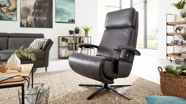 Ein modernes Wohnzimmer mit dem Interliving Sessel Serie 4541 - Relaxsessel F in grauem Leder auf einem Webteppich, umgeben von Pflanzen, Regalen, Sofa und von der Natur inspirierter Wandkunst. Durch die großen Fenster strömt Sonnenlicht herein.