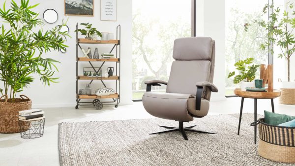 Ein modernes Wohnzimmer mit dem Sessel Serie 4541 - Relaxsessel F von Interliving auf einem geflochtenen Teppich, gepaart mit Zimmerpflanzen, einem runden Beistelltisch aus Holz, einem Weidenkorb, einem Metalldekorregal und großen Fenstern, die für natürliches Licht sorgen.