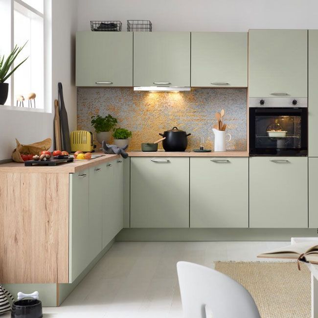 Eine moderne Küche mit hellgrünen Schränken, Holzakzenten, einem eingebauten Ofen und Arbeitsplattengeräten. Gemüse, Utensilien und Kochgeschirr stehen auf der Theke. Durch ein Fenster strömt Licht herein und erhellt den Raum.