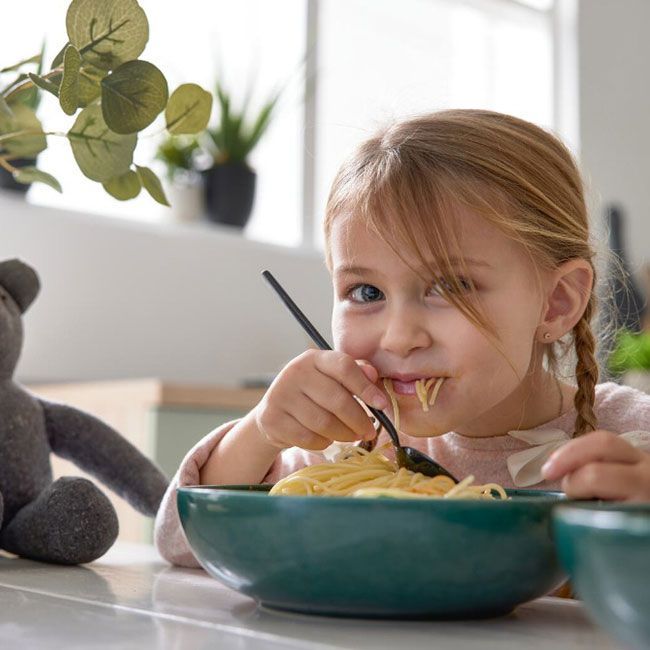 Ein junges Mädchen mit geflochtenem Haar sitzt an einem Tisch und isst lächelnd Spaghetti aus einer grünen Schüssel. Neben ihr liegt ein Stofftier, und im hellen Hintergrund sind Pflanzen zu sehen.