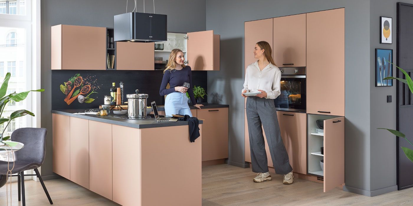 Zwei Frauen stehen und unterhalten sich in einer modernen Küche mit pfirsichfarbenen Schränken, schwarzen Arbeitsplatten und einer farbenfrohen Aufkantung mit Gemüsemotiven. Die eine hält eine Tasse, die andere hält Teller. Die Küche ist hell und stilvoll, mit Pflanzen und Wandkunst.